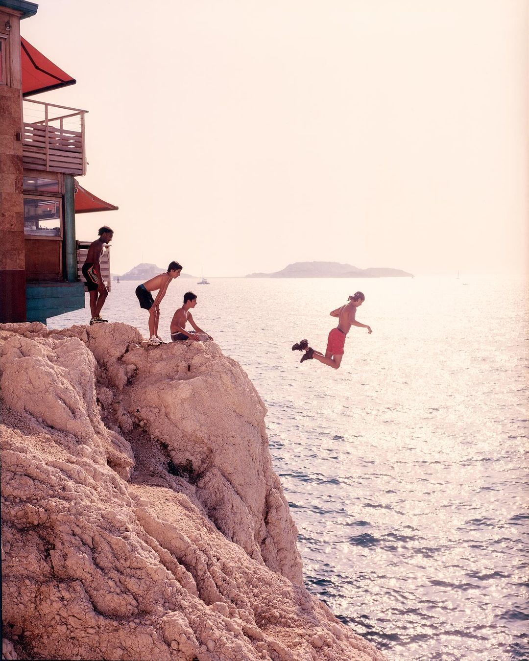 Four young men on a rocky cliff, one jumping into the ocean on a sunny day, building on the cliff, water, sky, rocks.