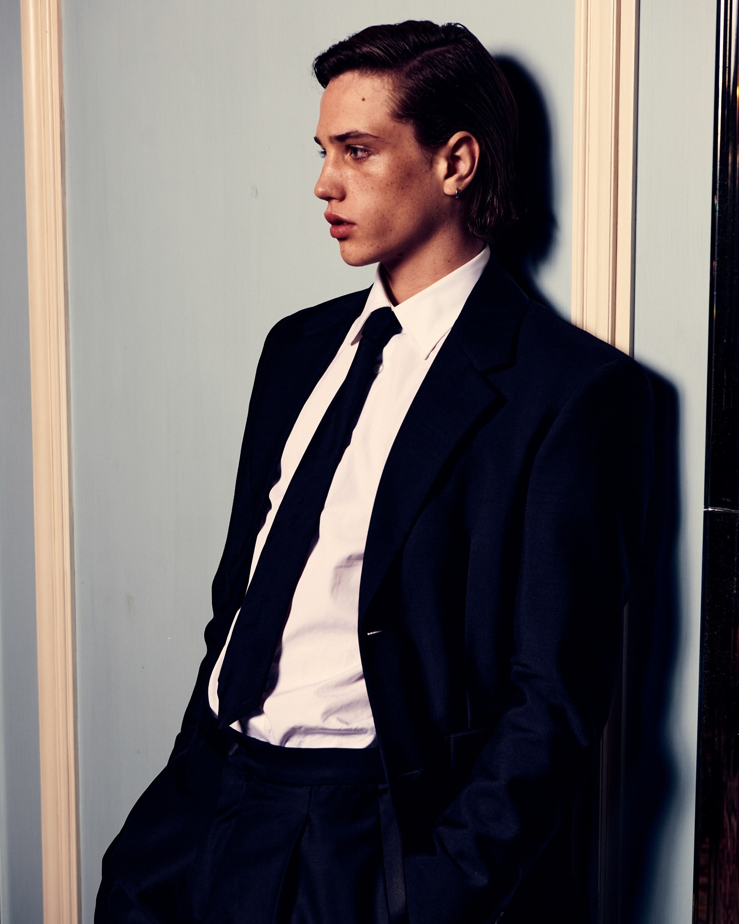 Portrait of a young adult man in a dark suit, white shirt, and dark tie leaning against a pale blue wall with white trim.