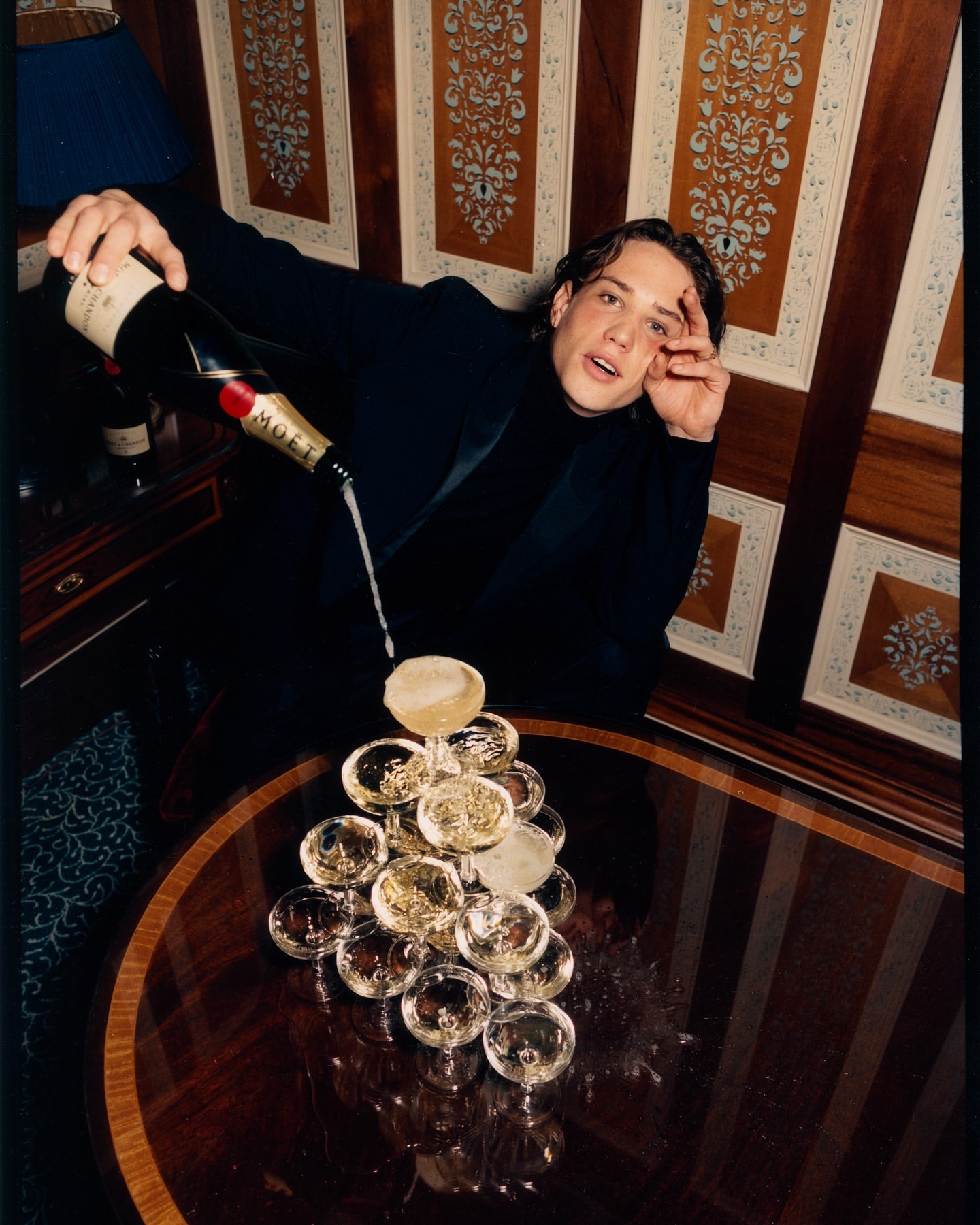 Man in black pours champagne over a tower of glasses indoors, with patterned wood paneling and warm lighting, creating a celebratory atmosphere.