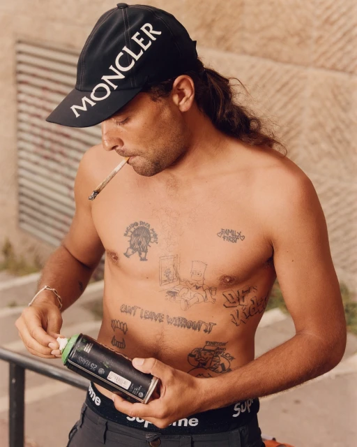 Shirtless man with a Moncler cap smoking while holding a spray paint can, posing outdoors with various tattoos on his chest and abdomen.