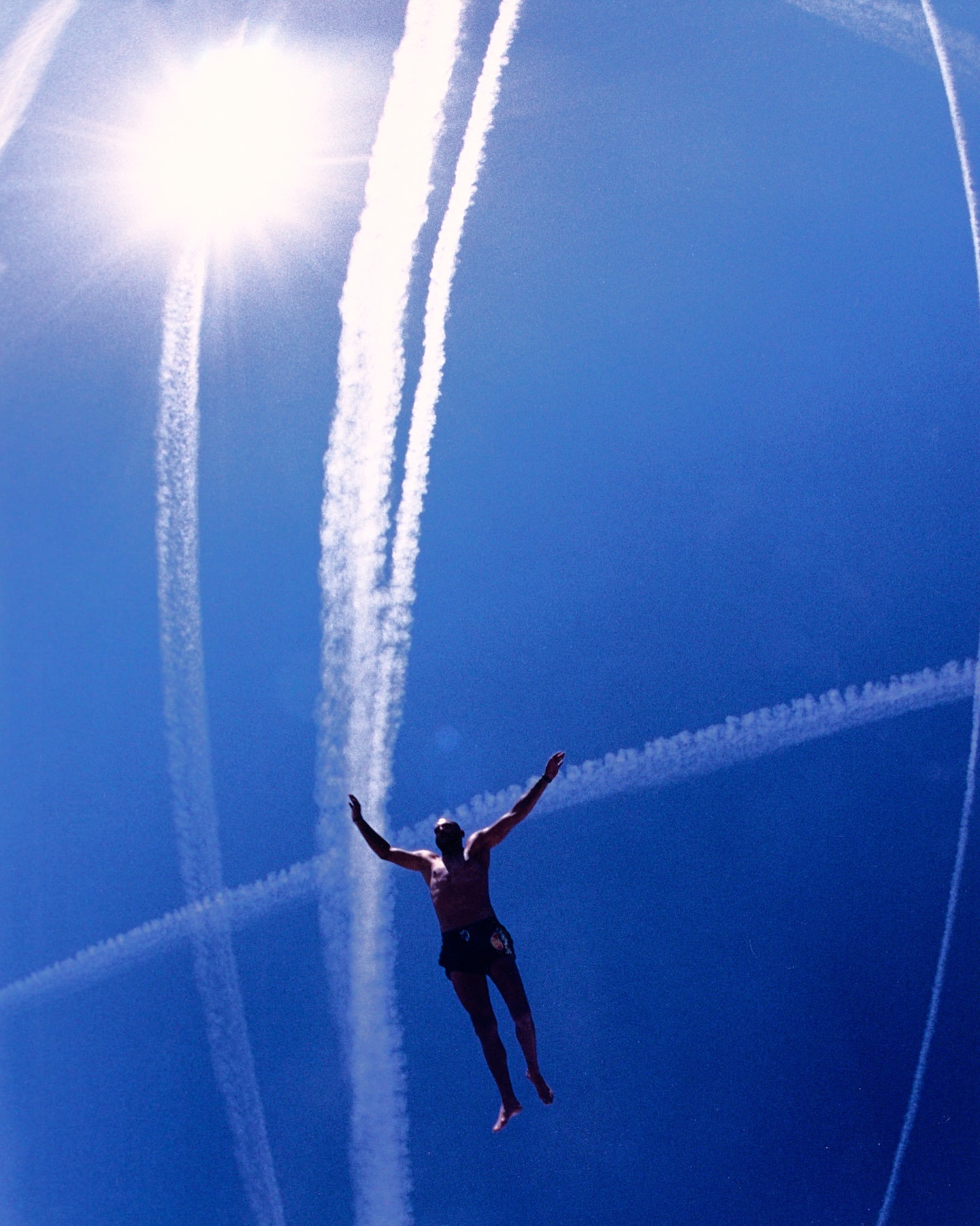 Man in freefall against bright blue sky with arms outstretched and contrails overhead, captured from a low angle.