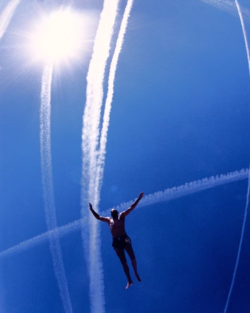 Man in freefall against bright blue sky with arms outstretched and contrails overhead, captured from a low angle.