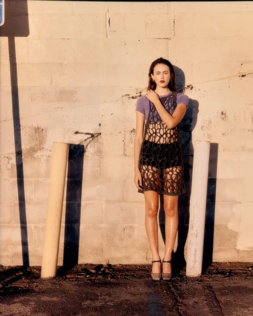 Young woman in crocheted dress poses against cinderblock wall, wearing platform heels, in an urban outdoor setting with bright sunlight.