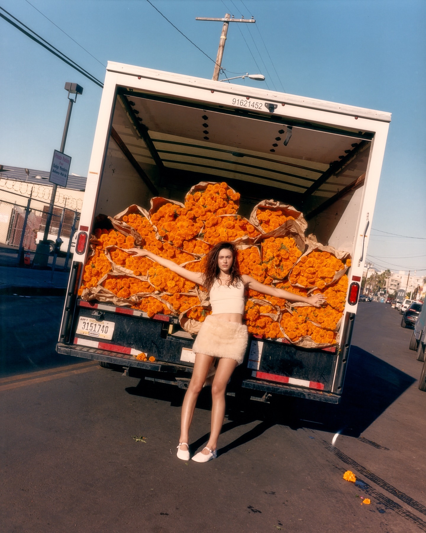 Young adult woman poses in front of flower-filled delivery truck on urban street under blue sky on sunny day. Fashion editorial.