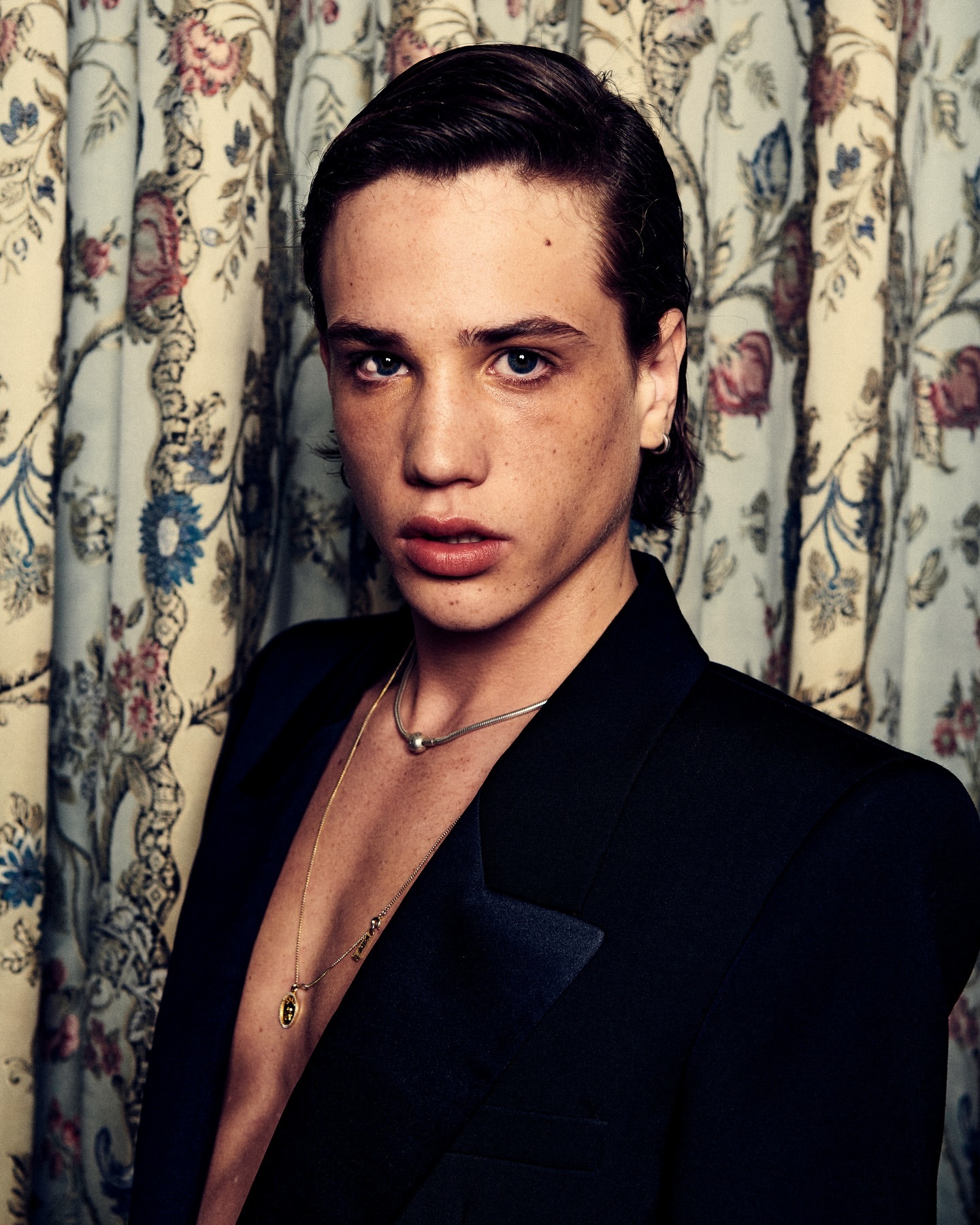 Close-up portrait of a young adult male in a black blazer, wearing necklaces, against a floral pattern backdrop in an indoor setting.