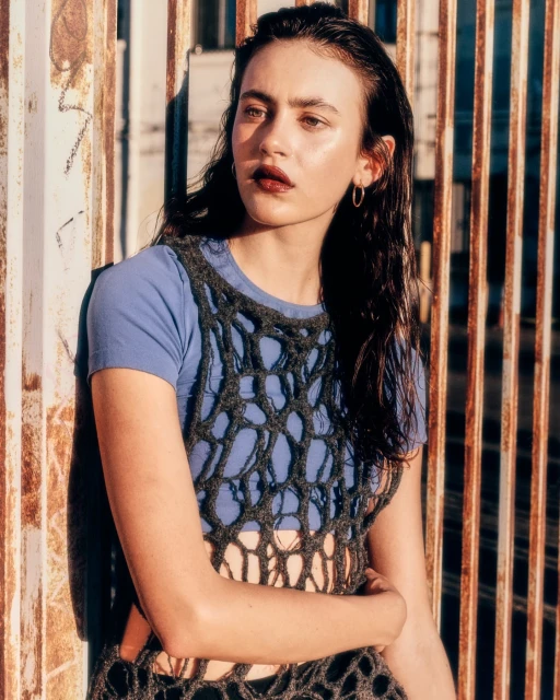 A young woman poses outdoors wearing a blue t-shirt and a grey crocheted top with a metal fence and textured wall in the background.