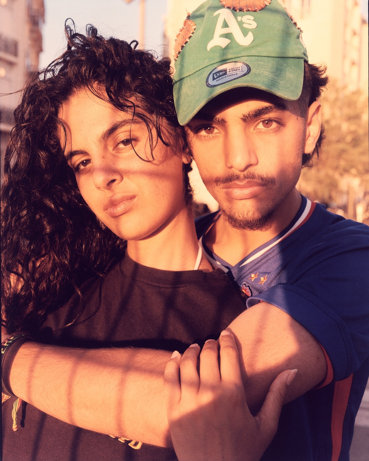 Warm, sunlit portrait of a young couple embracing outdoors, the woman with curly hair in a black top and the man in a baseball cap.