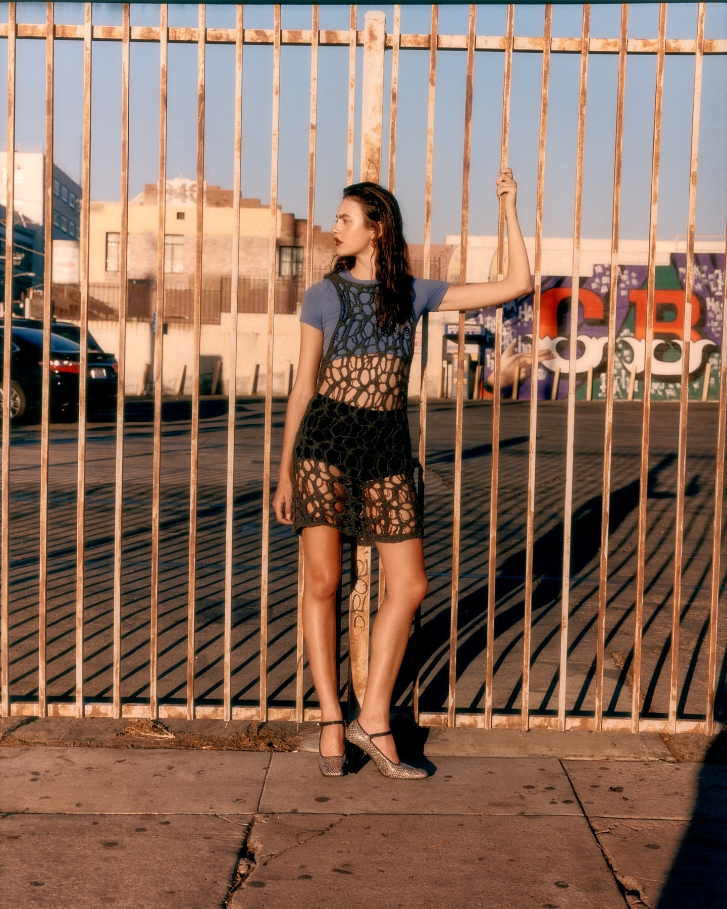 An adult female model poses in front of a metal fence in an urban setting, wearing a blue top, crochet dress, and metallic heels in sunlight.