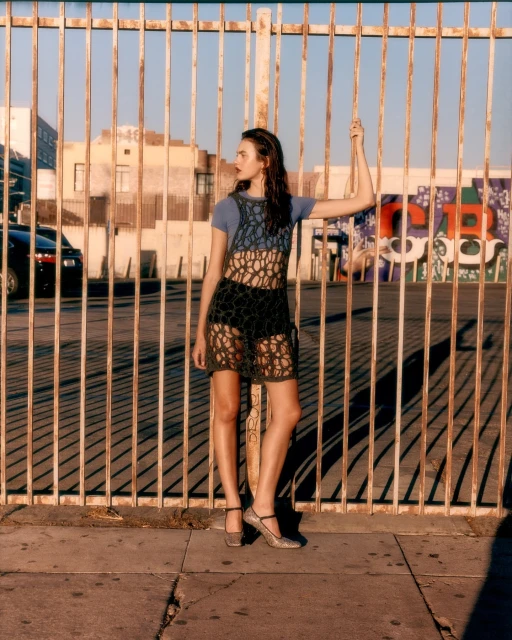 An adult female model poses in front of a metal fence in an urban setting, wearing a blue top, crochet dress, and metallic heels in sunlight.
