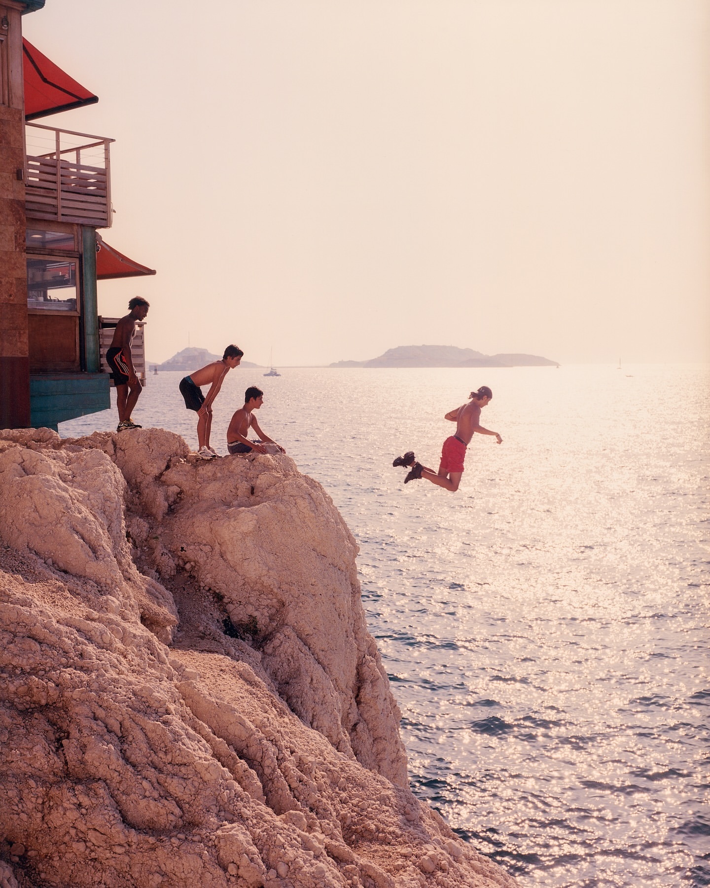 A group of young men on a rocky cliff, one jumping into the ocean. A building sits to the left, with an island in the distance.