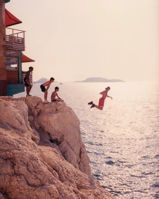 A group of young men on a rocky cliff, one jumping into the ocean. A building sits to the left, with an island in the distance.