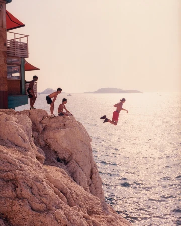 A group of young men on a rocky cliff, one jumping into the ocean. A building sits to the left, with an island in the distance.