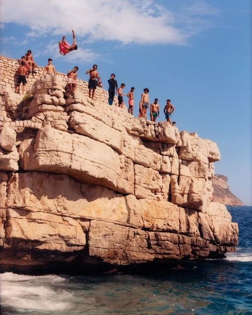A group of young men and boys on a rocky cliff by the ocean, one doing a backflip into the water under a sunny blue sky.