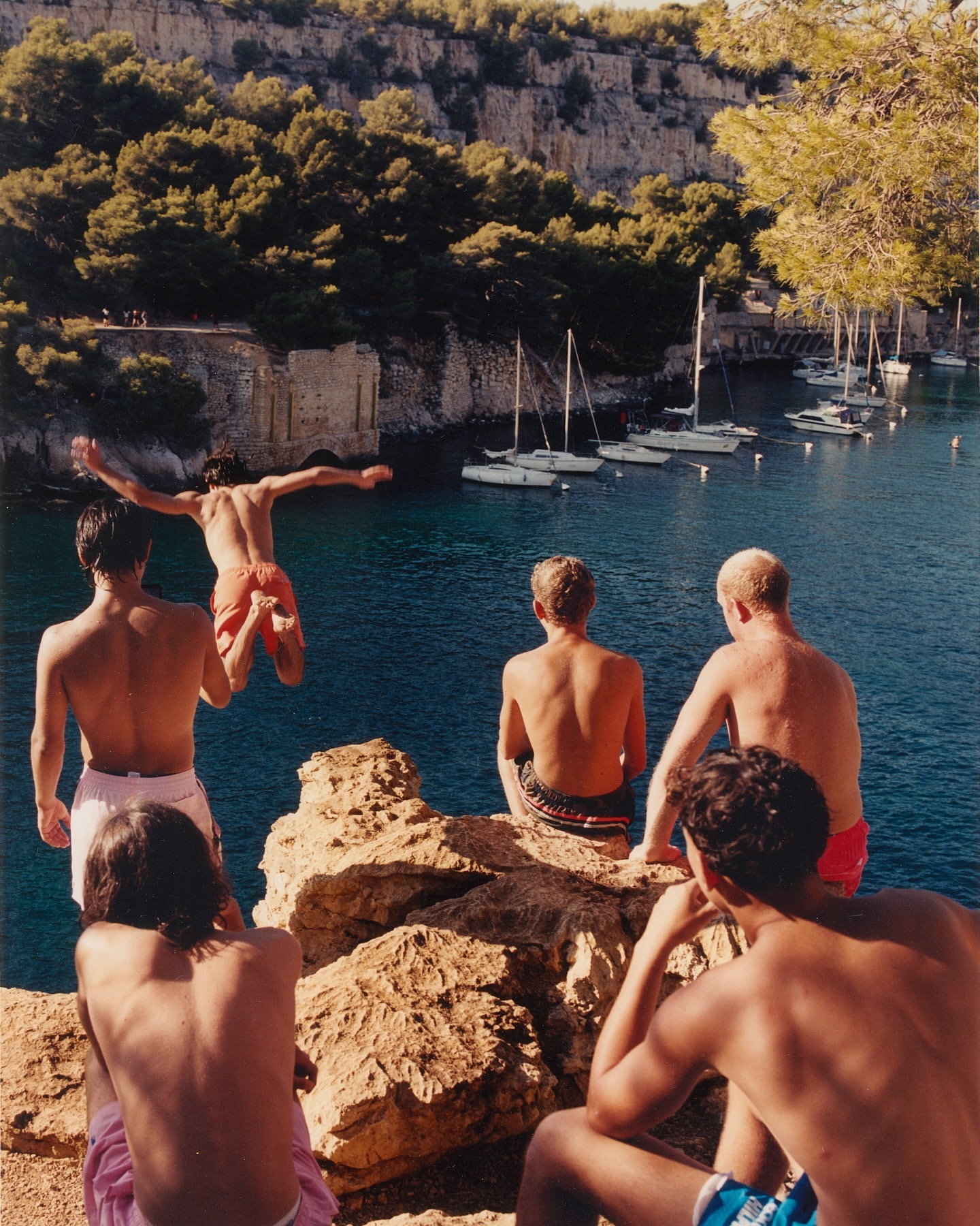 Group of shirtless young men on a rocky cliff edge; one dives into blue water. Sailboats visible in the background of an outdoor scene.