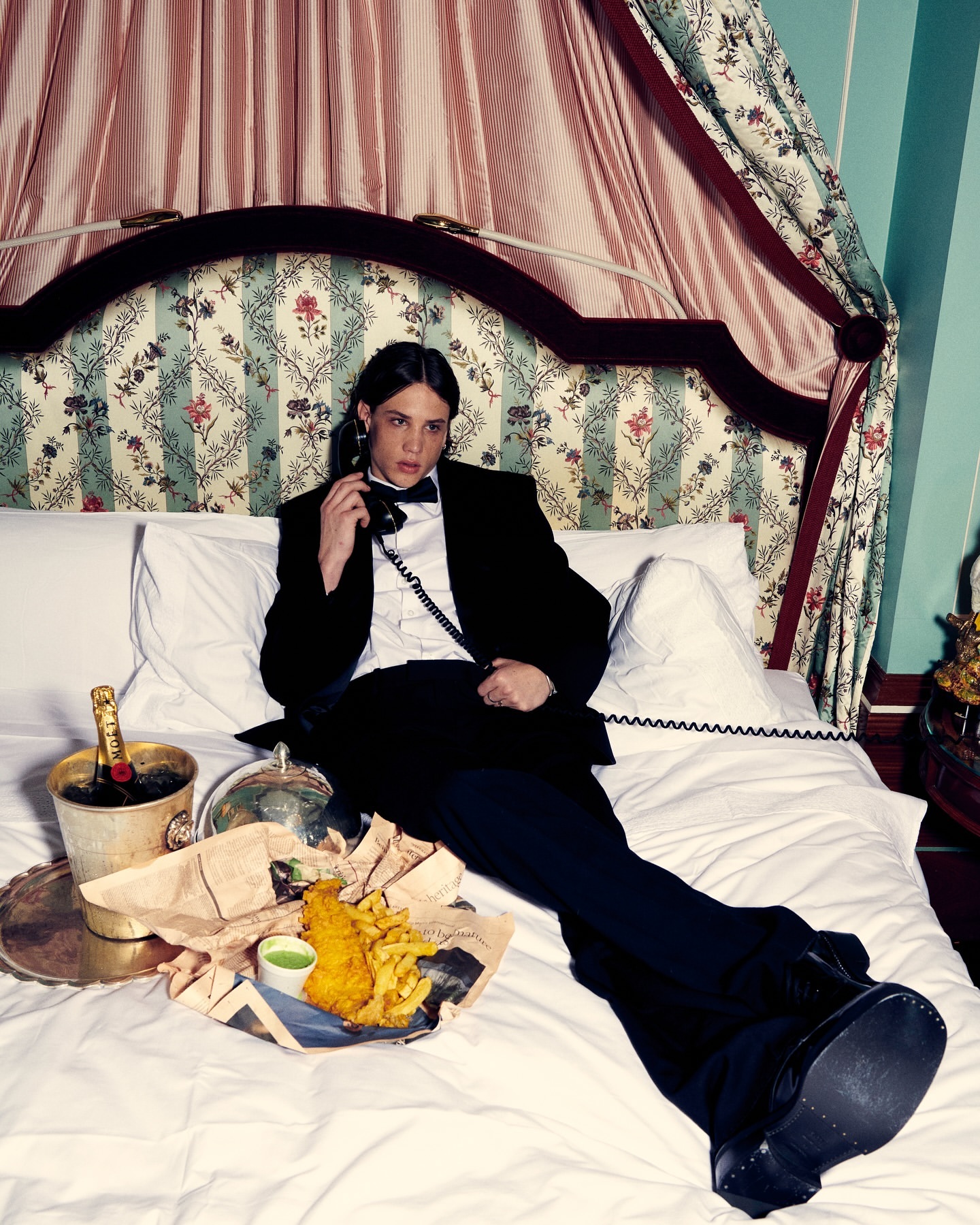 Adult male in a tuxedo lying on a bed, talking on the phone next to champagne in an ice bucket and fish and chips on a tray.
