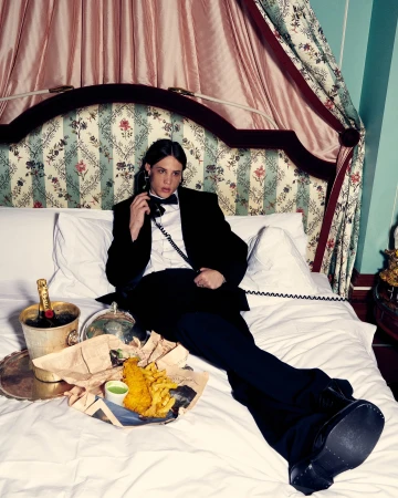 Adult male in a tuxedo lying on a bed, talking on the phone next to champagne in an ice bucket and fish and chips on a tray.