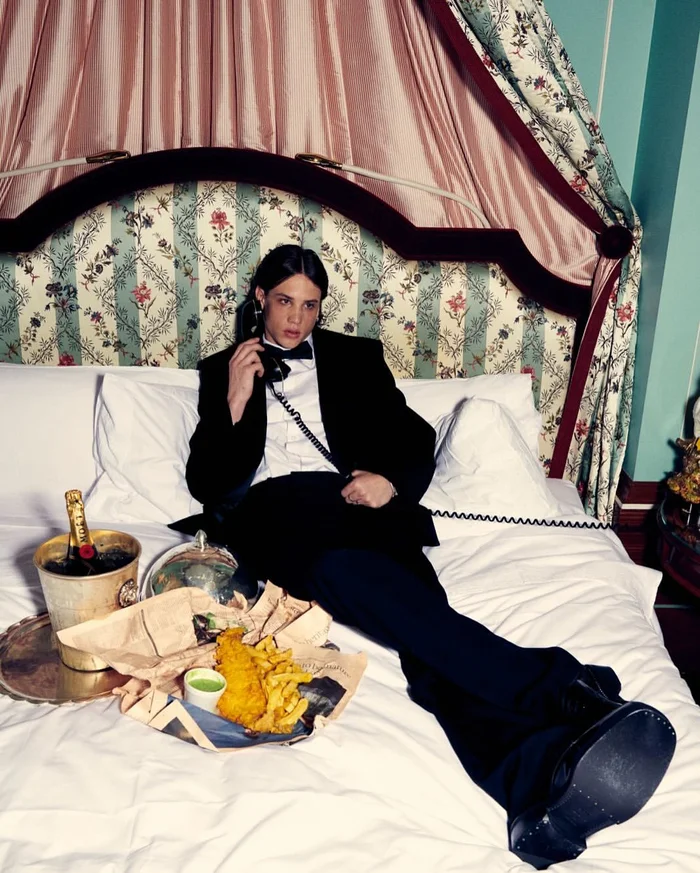 Man in a tuxedo holding a telephone receiver while seated on a hotel bed with fish and chips and champagne