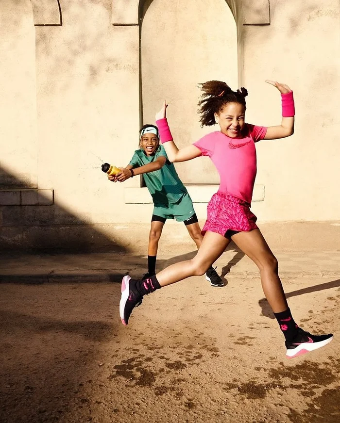 Two children playing outdoors in athletic wear, one leaping joyfully in pink while another squirts water in green.