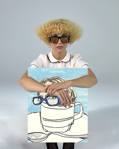 Woman with curly blonde hair and glasses holding an illustration of a person and coffee cup against a neutral gray background.