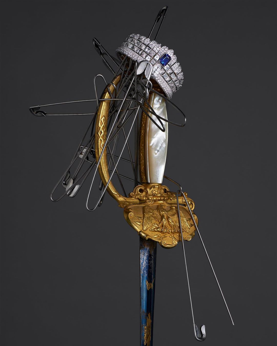 Ornate sword with a jeweled crown and pins. Decorative hilt with gold accents, pearl, and blue detailing. Studio lit on a dark background.