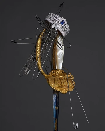 Ornate sword with a jeweled crown and pins. Decorative hilt with gold accents, pearl, and blue detailing. Studio lit on a dark background.