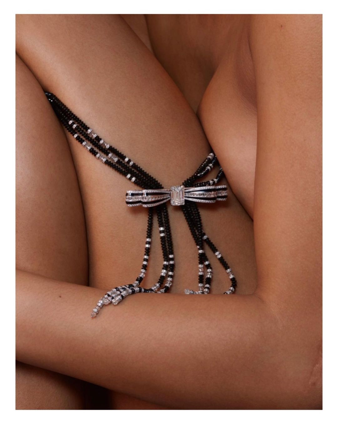 Closeup of a diamond bow necklace draped over a fair-skinned person's bare shoulder and arm, captured in soft, diffused lighting.