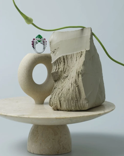 Close-up of an emerald and ruby ring on a marble pedestal next to a broken block against a light grey background with a plant stem.