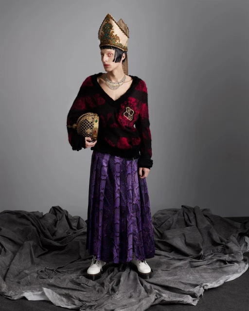A person in a high-fashion editorial-style portrait, wearing a distinctive mitre, ornate jewelry, patterned sweater and skirt, and holding a purse.
