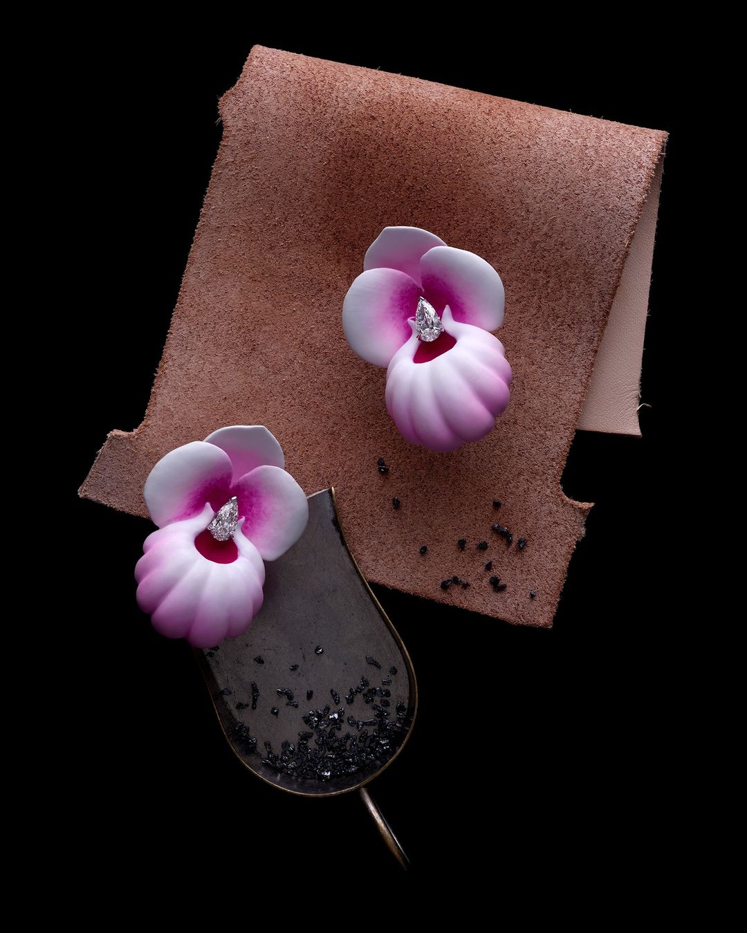 Overhead shot of two handcrafted earrings with floral design, set on a tan leather swatch, against a solid black background.