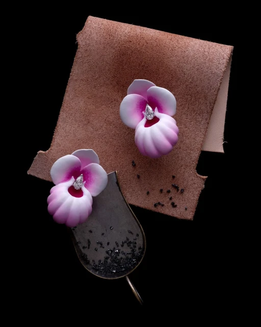 Overhead shot of two handcrafted earrings with floral design, set on a tan leather swatch, against a solid black background.