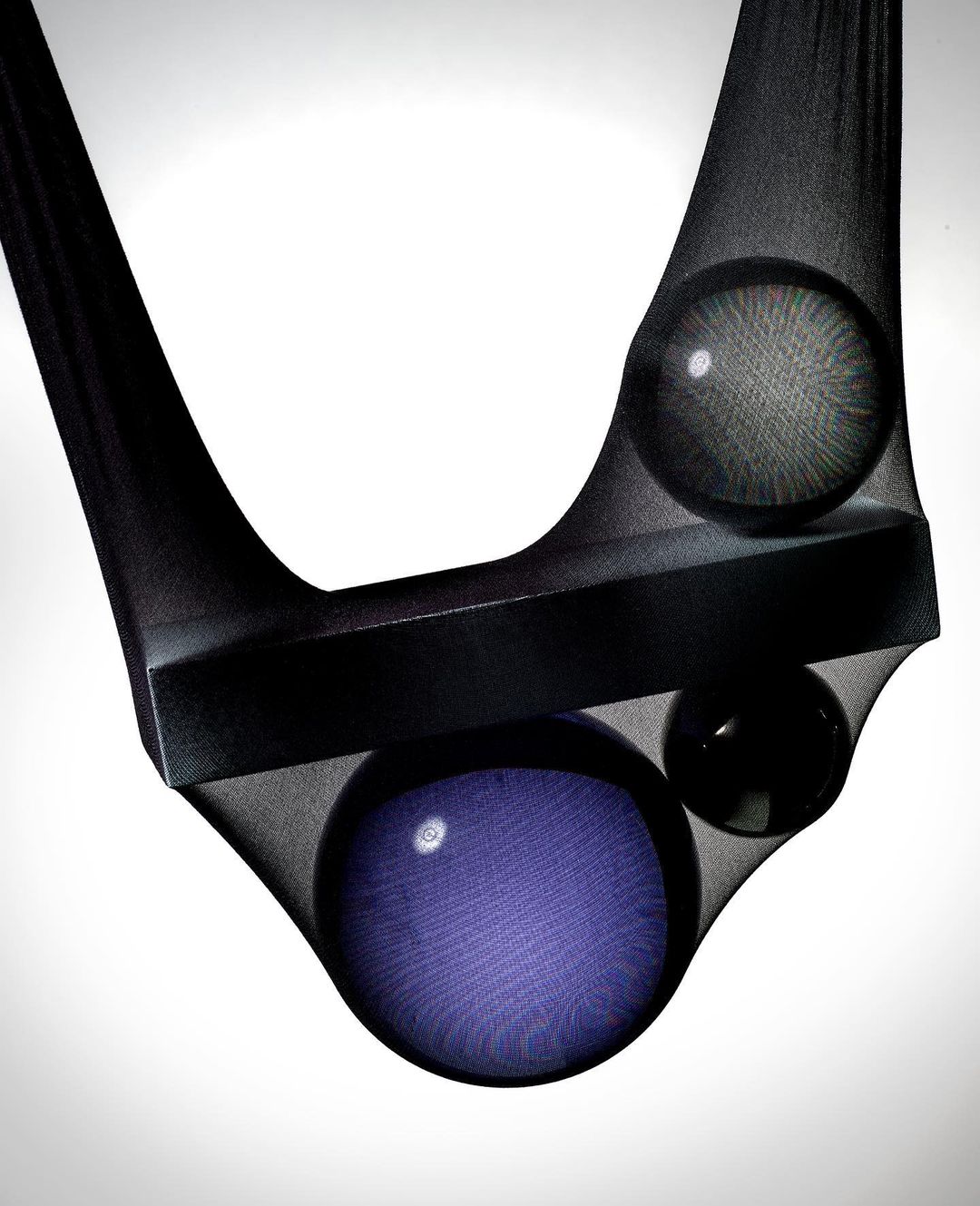 Abstract image with a black bar and three spheres suspended in dark sheer hosiery against a gradient white background.