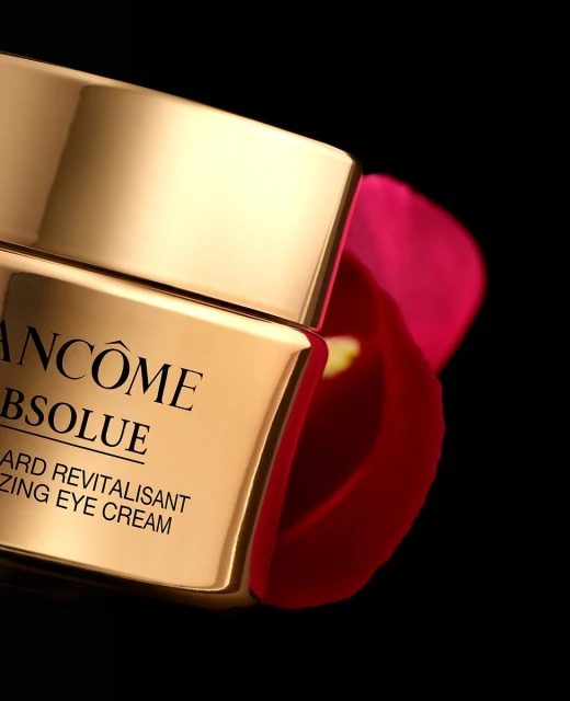 Close-up of gold Lancôme Absolue eye cream jar with black text, positioned near a soft-focus pink floral element against a black background.