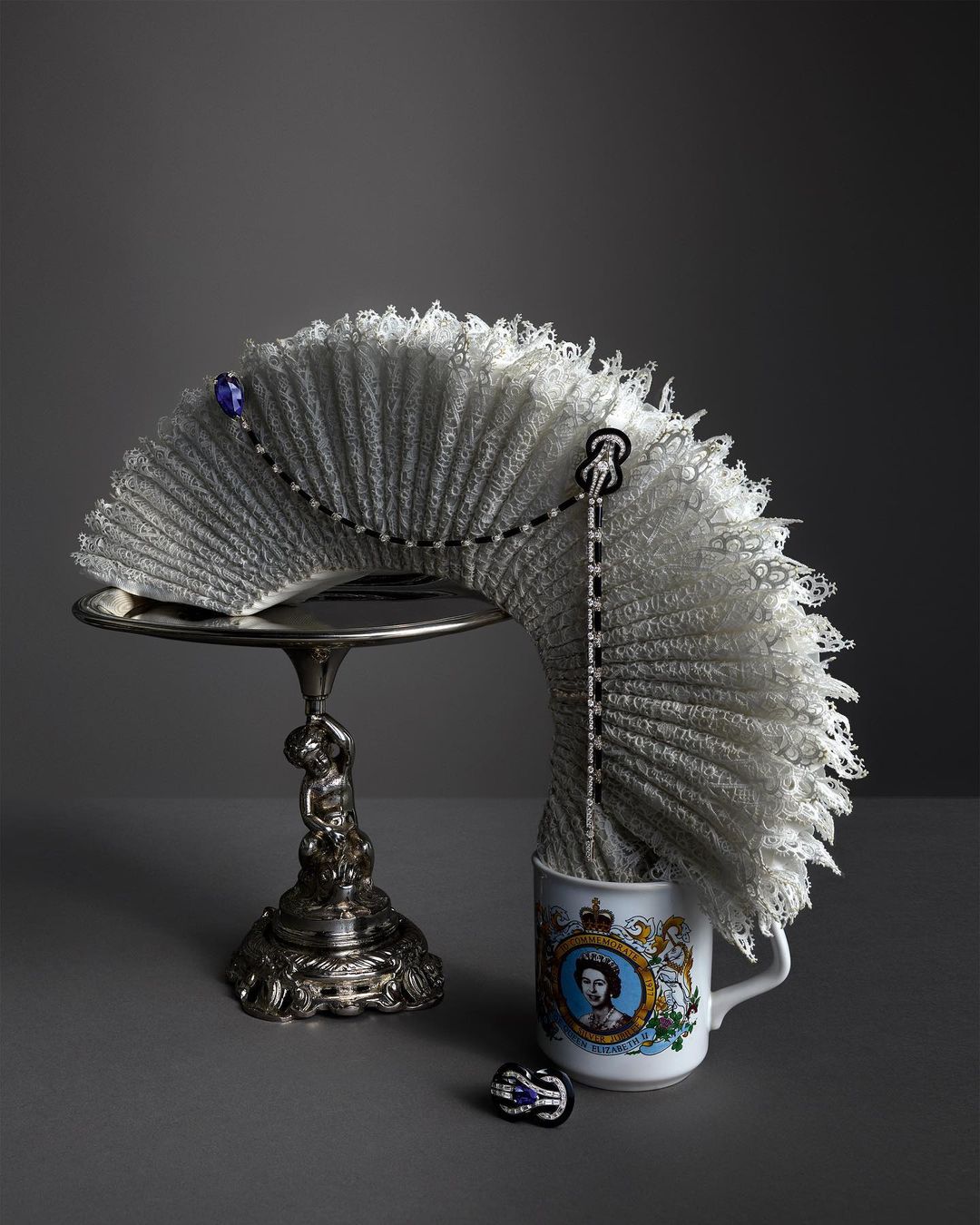 Lace fan on an ornate silver stand next to a mug with a portrait of Queen Elizabeth, set against a solid grey background.