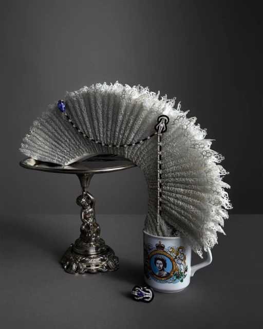 Lace fan on an ornate silver stand next to a mug with a portrait of Queen Elizabeth, set against a solid grey background.
