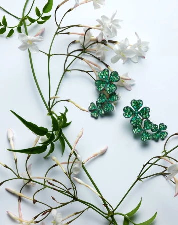 Close-up of green, sparkling clover-shaped jewelry interspersed with white jasmine flowers and vibrant green foliage on a white backdrop.