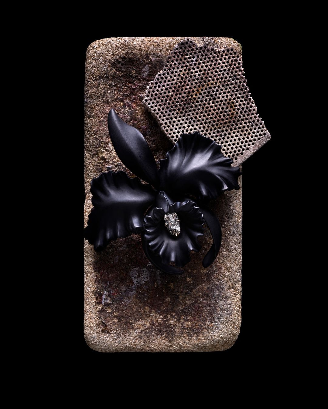 Still life shot of a black orchid jewelry piece on a stone surface, alongside a piece of patterned mesh, set against a dark background.