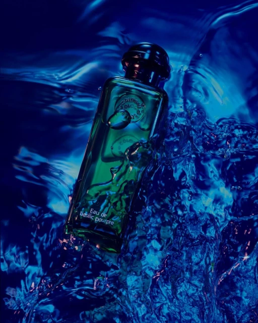 Close-up of a green Eau de Basilic Pourpre perfume bottle surrounded by splashing blue water in a product photography shot.