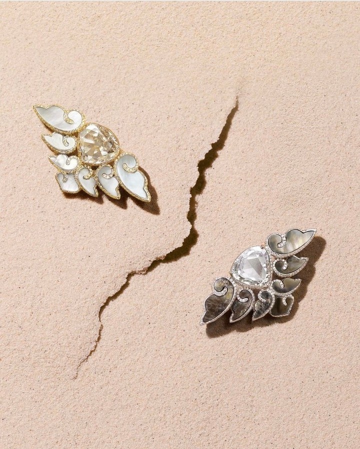 Two ornate gemstone earrings with pearl-like details rest on textured beige sand. Jewellery product photography showcases the intricate design.