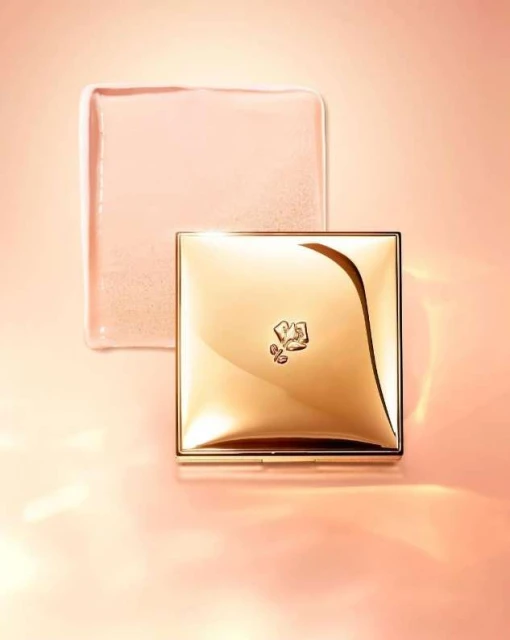 Golden Lancôme compact and translucent cube sit on a soft peach background with soft lighting. Beauty and luxury product still life.