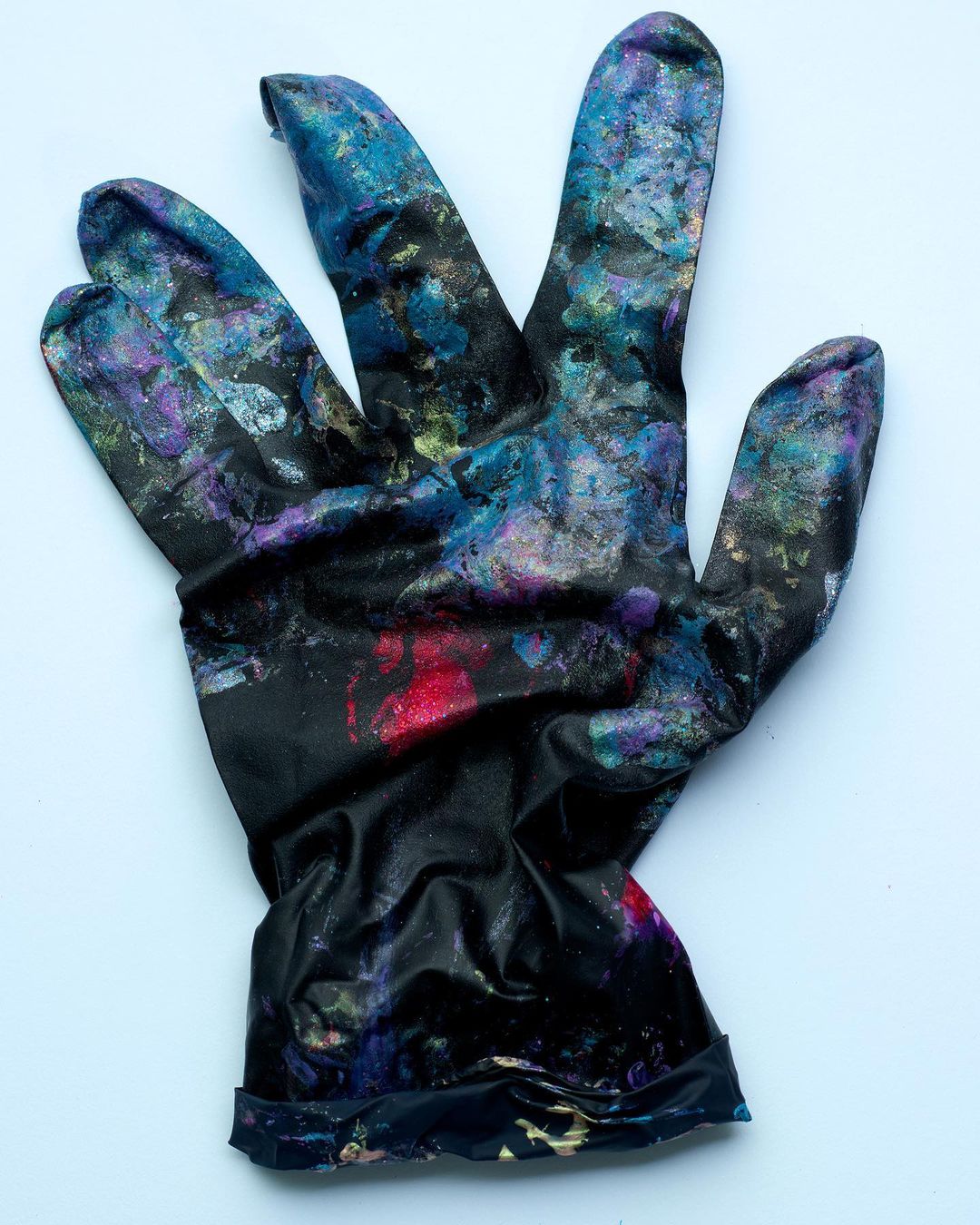 Close-up studio shot of a single used, black latex glove covered with paint splatters and placed against a white background.
