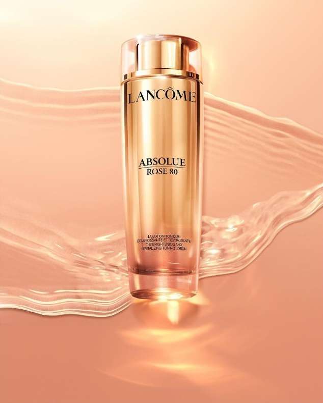 Lancome Absolue Rose 80 skin toner in gold bottle against peach backdrop with transparent water-like accents creating a soft, luxurious feel.