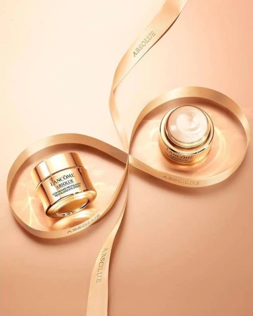 Two jars of Lancôme Absolue skincare product sit nestled within a spiraling ribbon against a light beige backdrop, studio product shot.