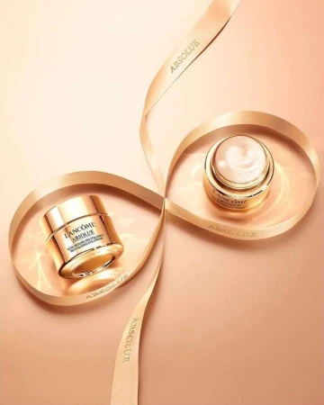 Two jars of Lancôme Absolue skincare product sit nestled within a spiraling ribbon against a light beige backdrop, studio product shot.