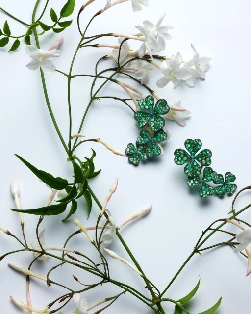 Close-up still life of green clover jewelry and white jasmine flowers on a white surface, showcasing jewelry and botanical elements.