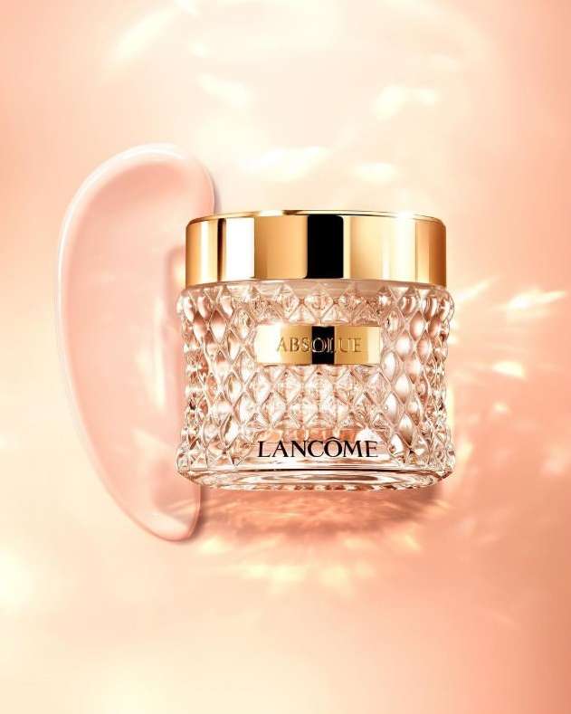 Close-up studio shot of Lancôme Absolue cream in a faceted glass jar with gold lid next to a swirl of pink cream on a peach background.