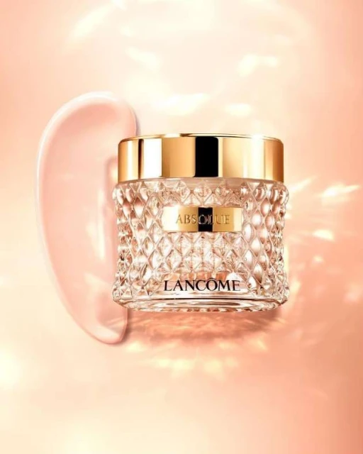 Close-up studio shot of Lancôme Absolue cream in a faceted glass jar with gold lid next to a swirl of pink cream on a peach background.