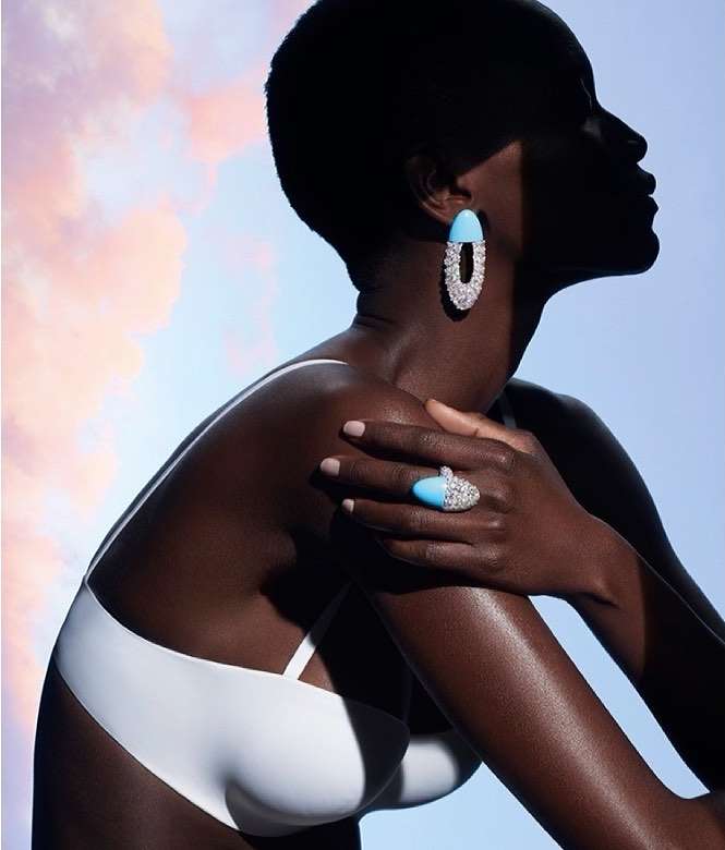 Elegant dark-skinned woman poses wearing a white bikini top and statement jewelry against a blue and pink pastel sky backdrop.