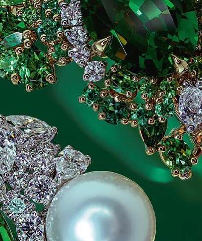 Close-up of jewelry featuring a large pearl, an emerald, and diamond accents on a green background. The shot focuses on detail and elegance.