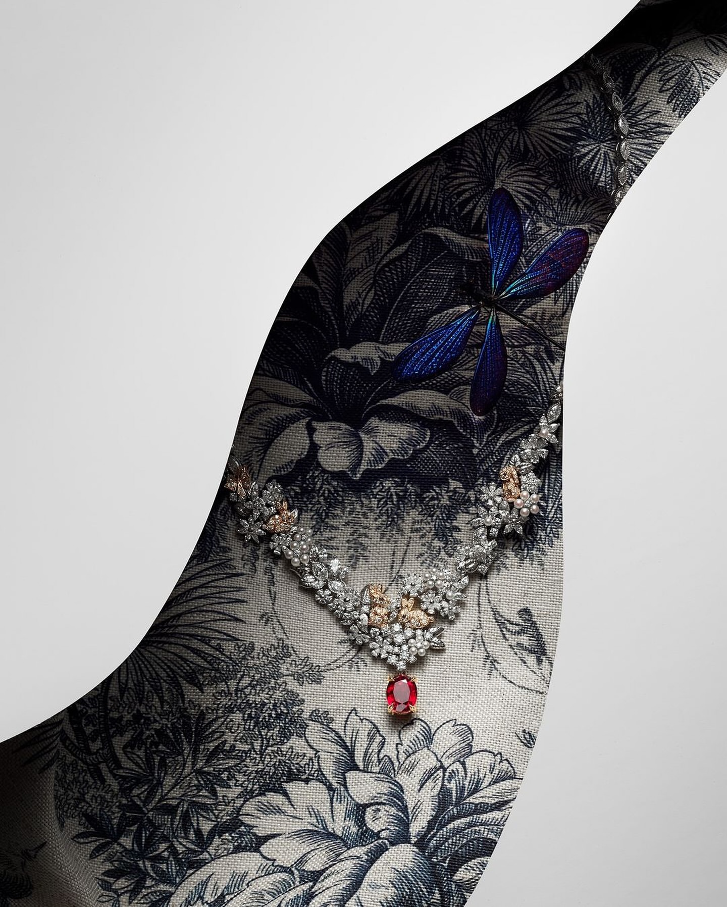 Elegant diamond necklace with a ruby pendant displayed against a botanical toile fabric and a gradient white backdrop in a jewellery advertisement.