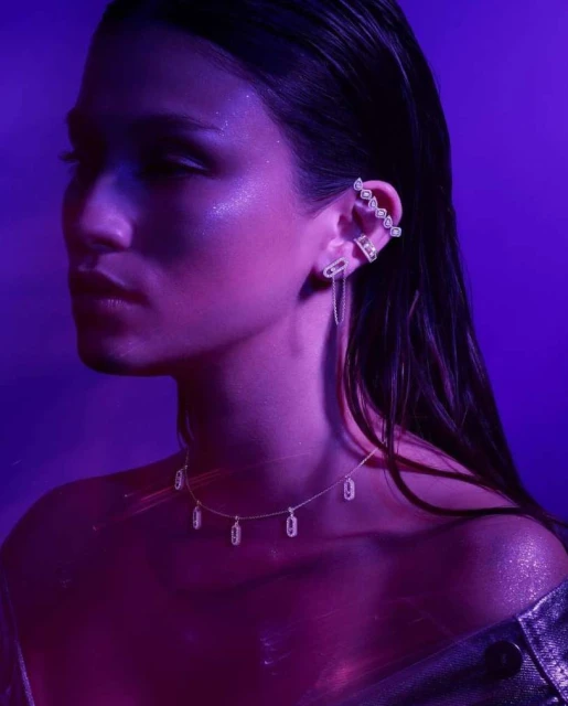 Close-up of a woman with multiple earrings and a diamond necklace, posing in a purple-lit indoor setting, beauty and fashion.